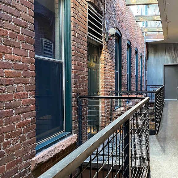 partially covered  long exterior hallway with brick walls and a metal railing.