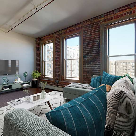 a living room with a grey couch, tv and coffee table. exposed brick walls have 3 large windows overlooking cityscape.