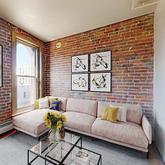 a living room with a sofa and coffee table, near exposed-brick wall with 2 large windows.