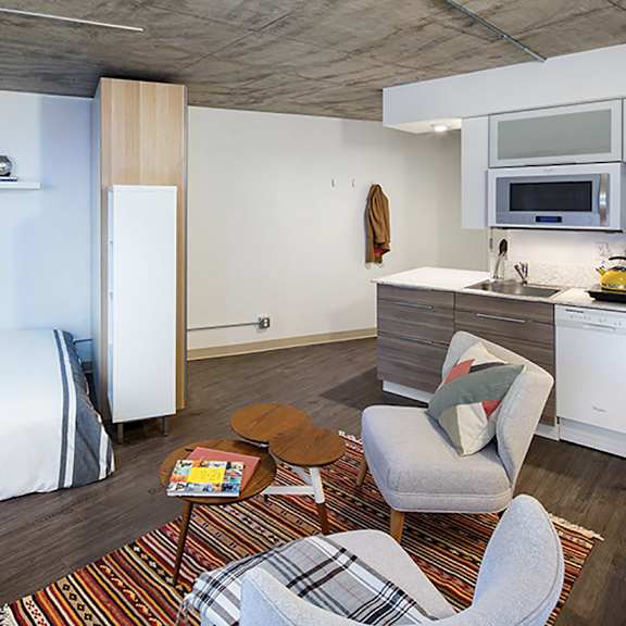 a bedroom with a bed, chair, and a kitchen area.