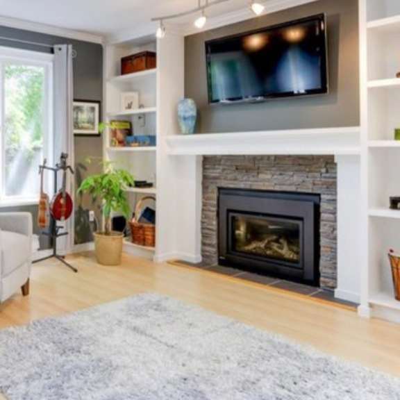 a living room with a grey couch, a grey chair, a white fireplace, a grey rug, a clock on the wall, a television on the wall, and a window with curtains.