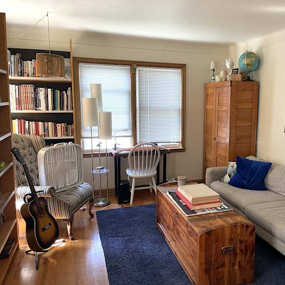 a living room with a grey couch, a wooden bookshelf, a chair, a guitar, a cabinet, a table, a window with blinds, a rug, a blue pillow, a picture frame, and a globe.