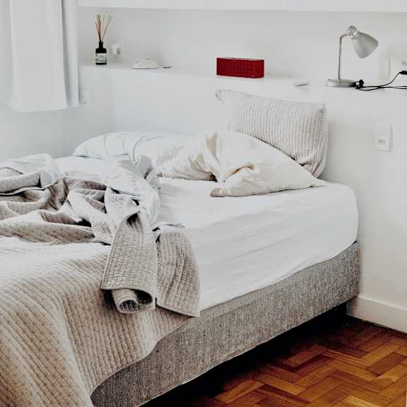 a white bed with a grey blanket and pillow with a white lamp on the side table.