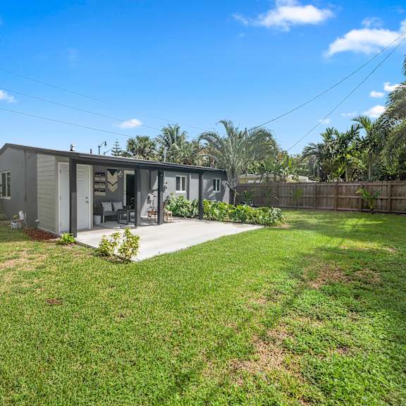 a house with a grey exterior is surrounded by a green lawn and palm trees.