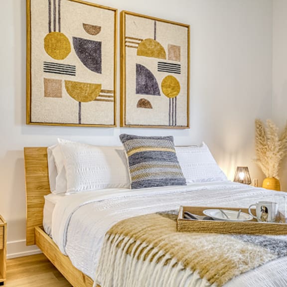 a bedroom with a bed, pillows, a tray with glasses and a phone on it, a nightstand, and two framed artworks on the wall.