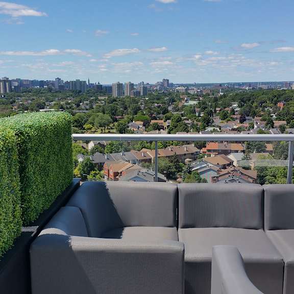 a grey couch with a white table in front of a green hedge and a cityscape in the background.