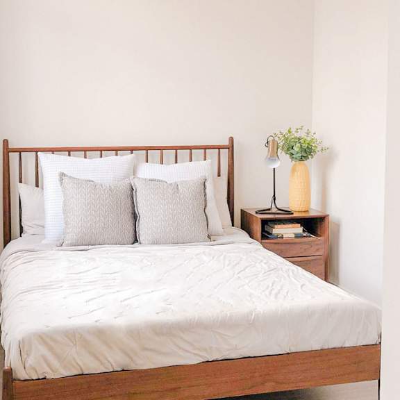 a bed with a white comforter and four pillows sits in a room with a small wooden nightstand holding a vase with flowers and a lamp.