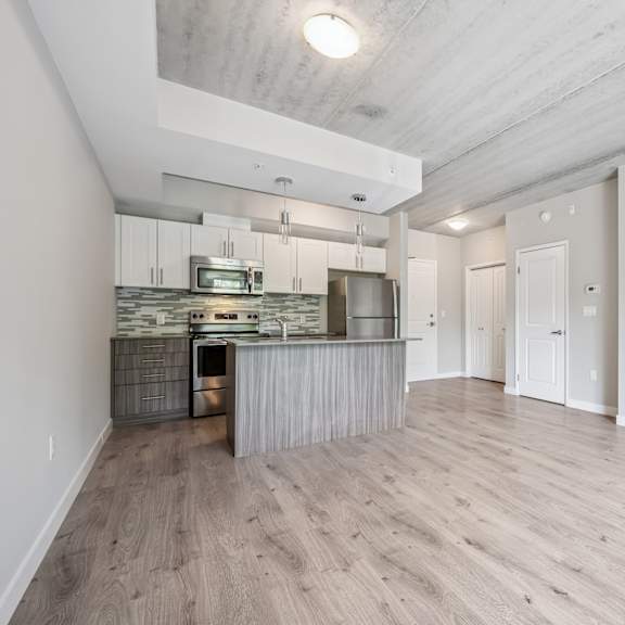 a spacious, empty room with wooden flooring and a kitchen area in the background.