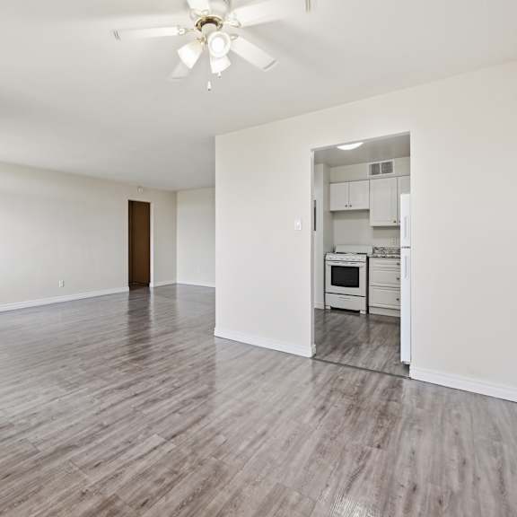 a spacious room with a ceiling fan and light, hardwood floors, and a kitchenette in the background.