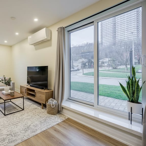 a living room with a grey couch, a coffee table, a flat screen tv, and a large window with a view of a green lawn.