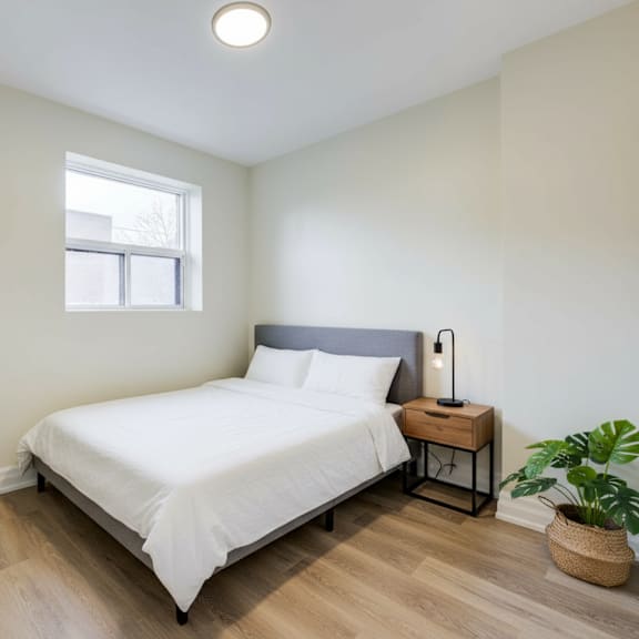 a bedroom with a white bed, a small table with a lamp, and two potted plants.
