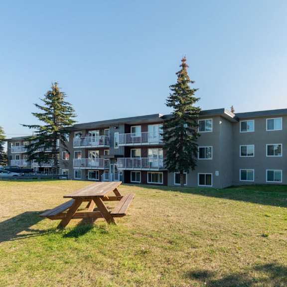 a wooden picnic table is in the foreground of a grassy area in front of apartment buildings.