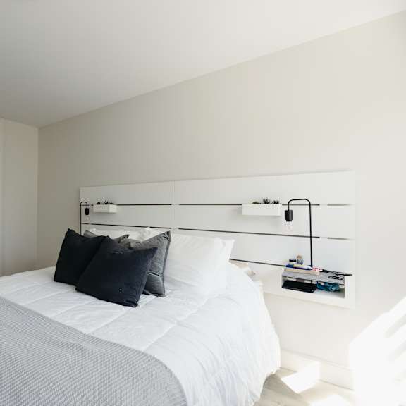 a white bed with a white headboard and a white nightstand with a lamp on it.