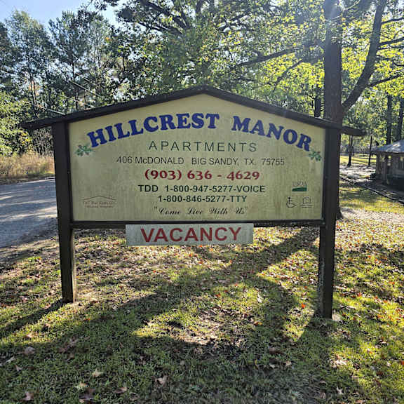 a sign for hillcrest manor apartments is displayed in front of a wooded area.