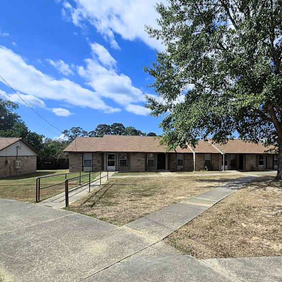 a large tree stands in front of a building with a fence and a grassy area in the foreground.