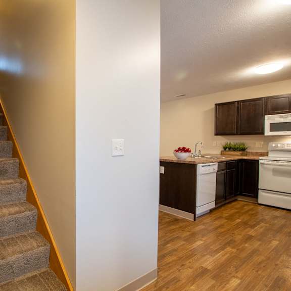 a staircase with a carpeted runner and wooden handrail leads to a kitchen with dark wood floors and white appliances.