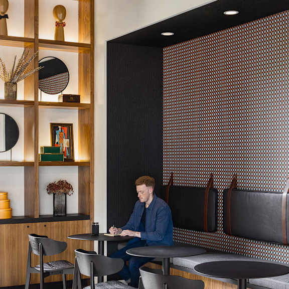a man is sitting at a table in a room with a wooden shelf and a black and white patterned wall.