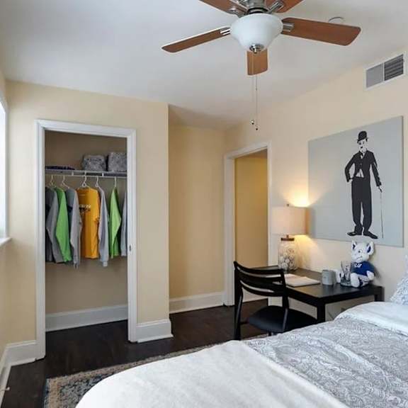 a bedroom with a bed, a ceiling fan, a chair, a table, a potted plant, and a painting of a person.