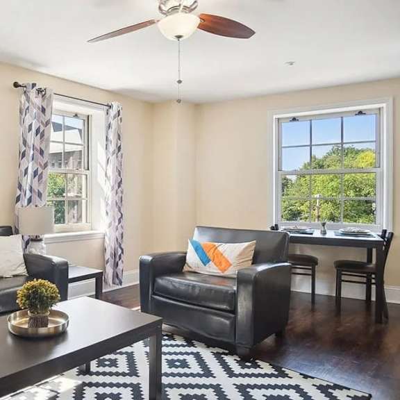 a black leather couch sits in a living room with a ceiling fan and a window with a view of trees.