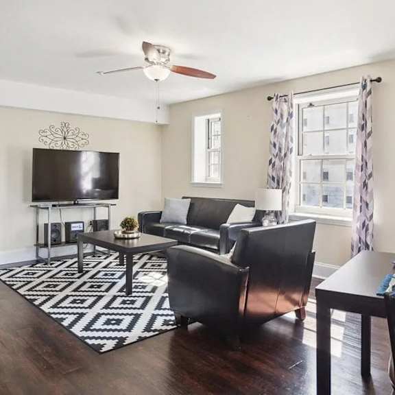 a living room with a black and white rug, a black couch, a black chair, a black coffee table, a flat screen tv, and a window with curtains.