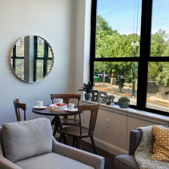 a living room with a grey couch, a wooden bookshelf, a round mirror, and a window with a view of trees.