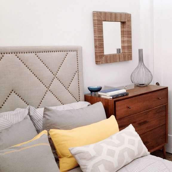 a bed with a grey headboard and pillows in front of a mirror and a dresser.