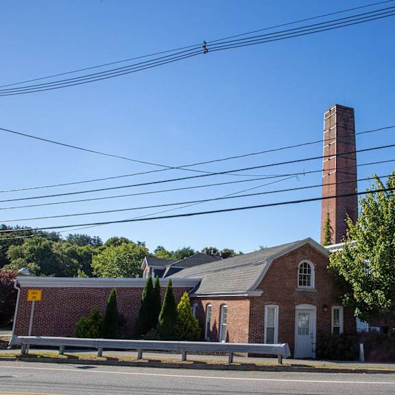 a brick building with a tall chimney is surrounded by trees and parked cars.