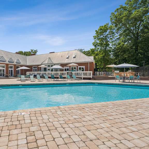 a large outdoor swimming pool with a brick patio and a house in the background. at the timbers apartments, virginia
