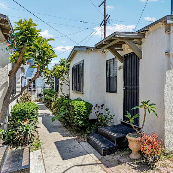 a small white house with a black door and windows surrounded by plants and a brick walkway.