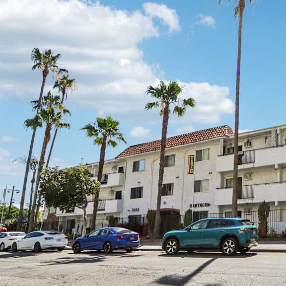 cars parked in front of a white apartment building with palm trees.
