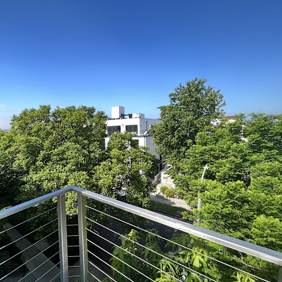Thumbnail 2 of 7 A balcony with a metal railing overlooks a lush green area with trees and a white building in the distance.