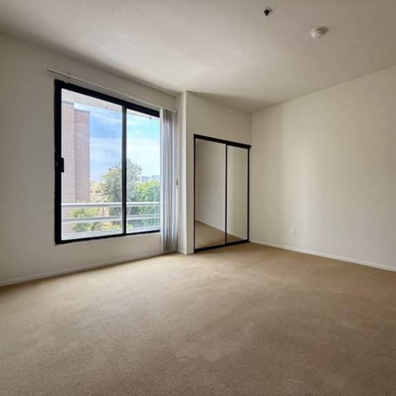 Thumbnail 20 of 27 A room with a carpeted floor, a window with a view of a building and trees, and a sliding door.