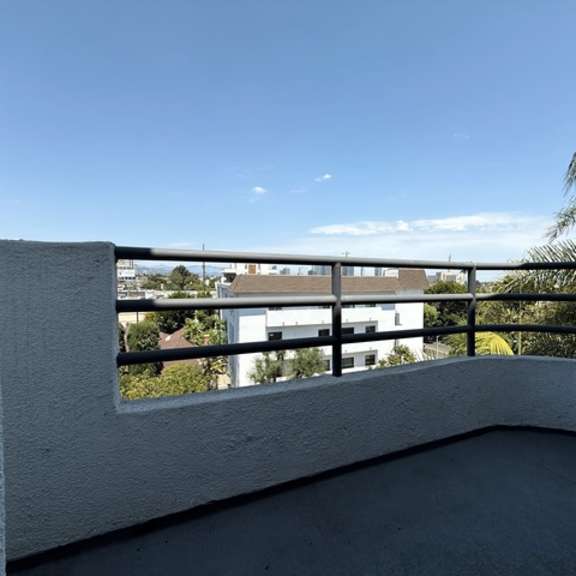 Thumbnail 20 of 28 A balcony with a white wall and a black railing overlooking a building and trees.