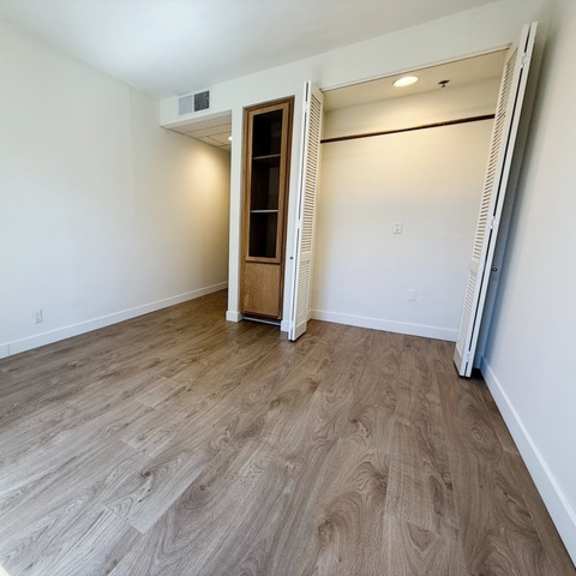 Thumbnail 5 of 13 A room with a wooden floor and a white wall with a doorway leading to another room.