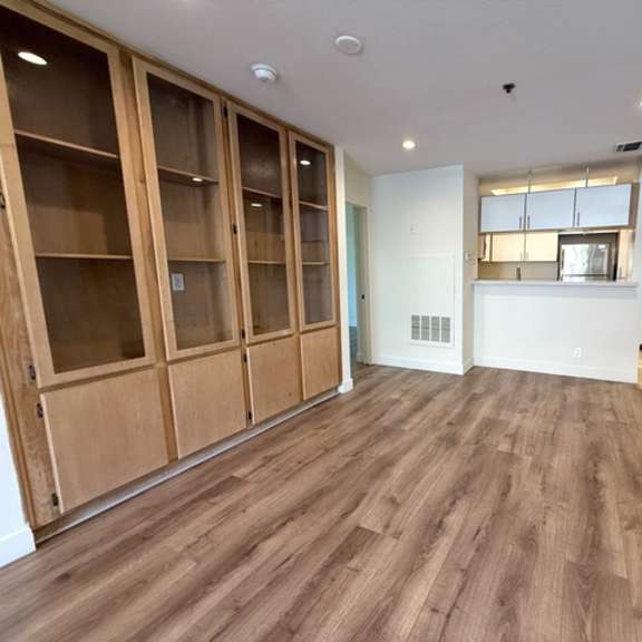 Thumbnail 10 of 44 A room with wooden floors and white walls, featuring a row of wooden shelves and a kitchenette in the background.