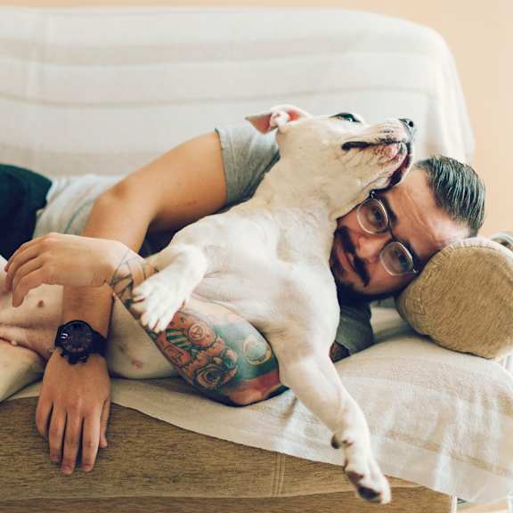 A man with tattoos and glasses is lying on a couch with a white dog on his back.