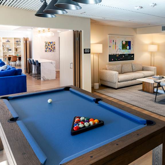 a pool table with a blue cloth and a rack of balls is in the foreground of a room with a couch, chairs, and a painting on the wall.