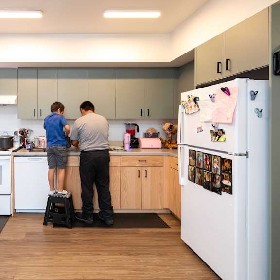 two men are standing in a kitchen with a refrigerator covered in magnets and pictures.