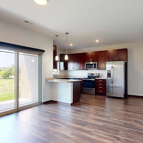 Thumbnail 13 of 29 An open living room with sliding glass door to a patio, and modern kitchen with dark wooden cabinets.