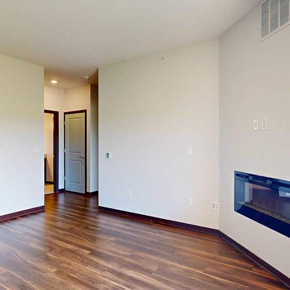 Thumbnail 19 of 29 A living room with hardwood flooring, an electric fireplace, and a hallway to a bathroom and bedroom.