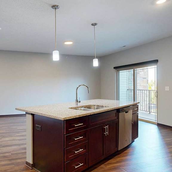 Thumbnail 5 of 34 An open-concept living room with hardwood flooring and a sliding glass door to a balcony, that connects to a modern kitchen with a large central island.