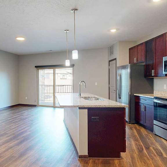 Thumbnail 10 of 34 An open-concept living room with hardwood flooring and a sliding glass door to a balcony, that connects to a modern kitchen with a large central island.