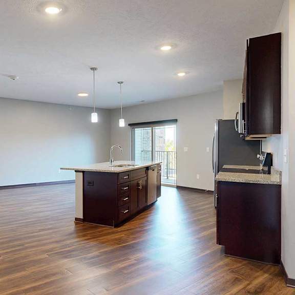 Thumbnail 4 of 34 An open-concept living room with hardwood flooring and a sliding glass door to a balcony, that connects to a modern kitchen with a large central island.