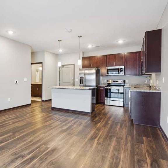 Thumbnail 10 of 15 A living room with hardwood-style flooring and kitchen with dark wooden cabinets.