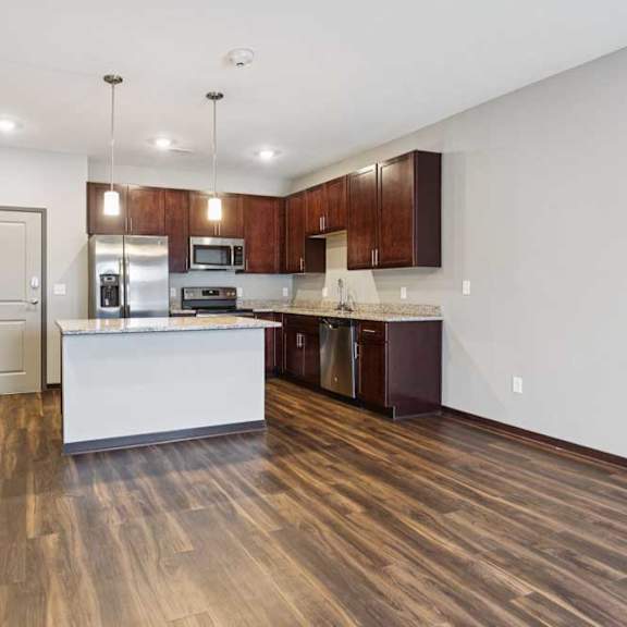 Thumbnail 9 of 15 A living room with hardwood-style flooring and kitchen with dark wooden cabinets.