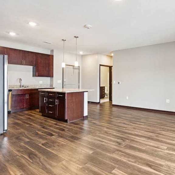 Thumbnail 12 of 18 A living room with hardwood-style flooring and kitchen with dark wooden cabinets.