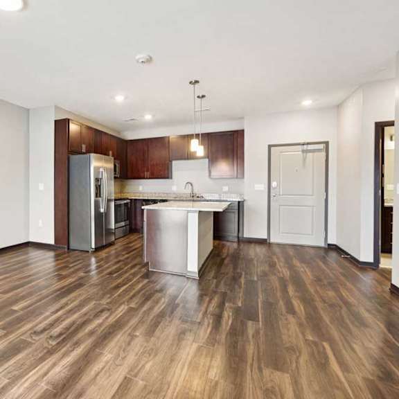 Thumbnail 3 of 18 A living room with hardwood-style flooring and kitchen with dark wooden cabinets.