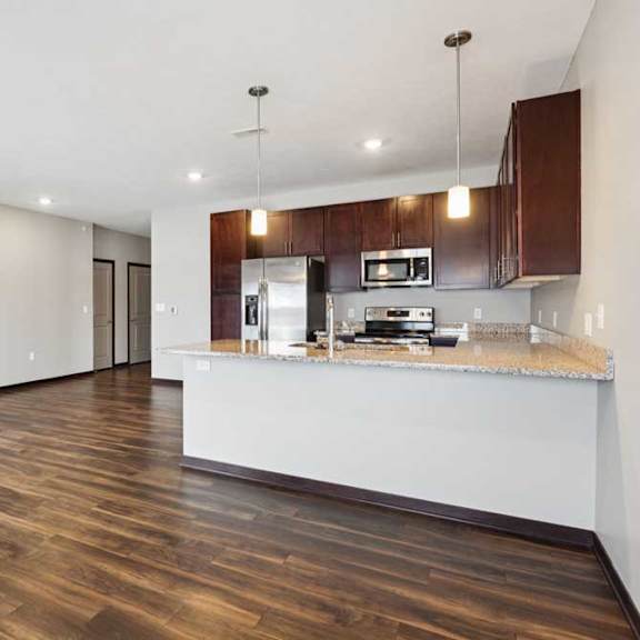 Thumbnail 3 of 19 A living room with hardwood-style flooring and kitchen with dark wooden cabinets.