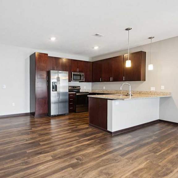 Thumbnail 4 of 19 A living room with hardwood-style flooring and kitchen with dark wooden cabinets.