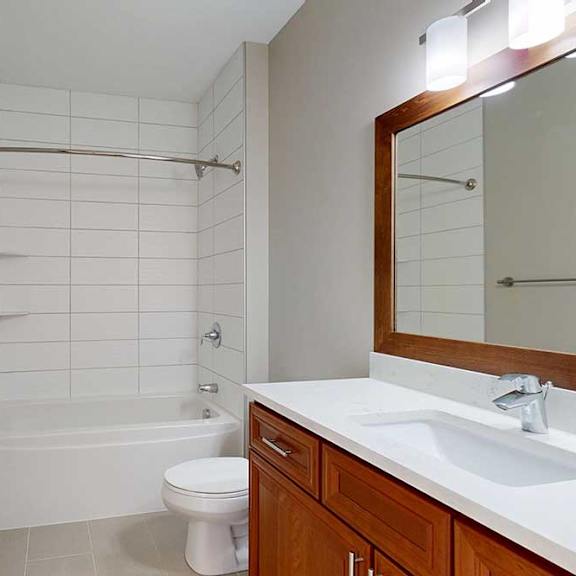 Thumbnail 27 of 27 A bathroom with cherry-stained wooden cabinets, white quartz vanity, and a tiled tub shower.